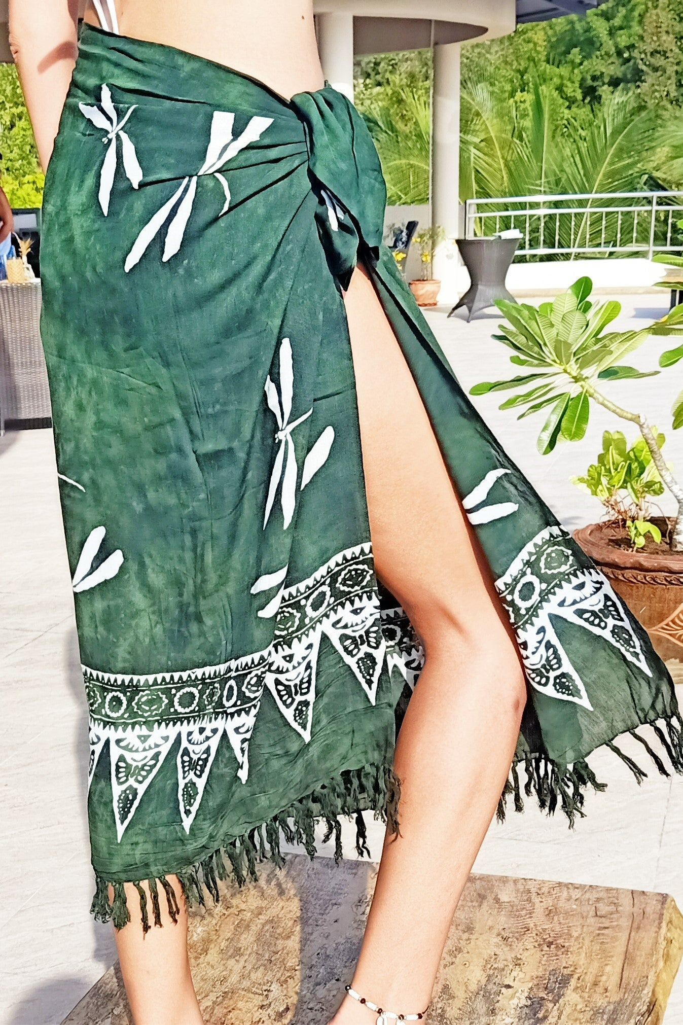 Woman wearing wrap around skirt with leg showing. Tied at waist with fringe at the bottom. Green background with white dragonfly pattern.