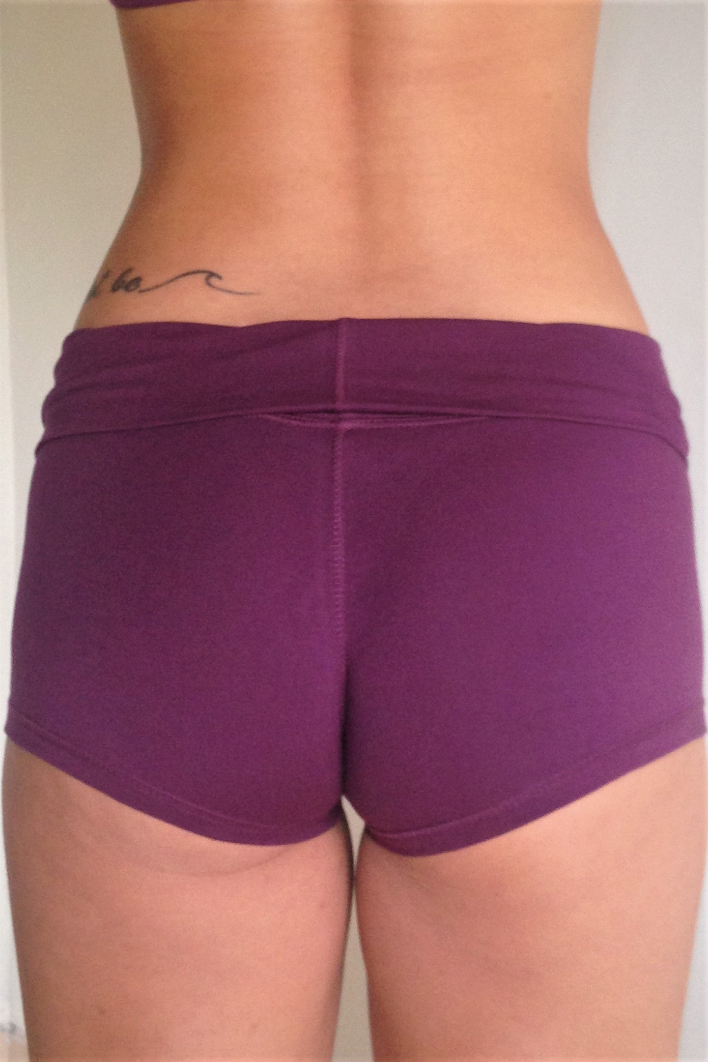Back view of solid Plum purple yoga shorts with fold over waist, can be worn higher waisted, or low on the hips. Small black Lotus Flower embroidery. 90% cotton 10% spandex.