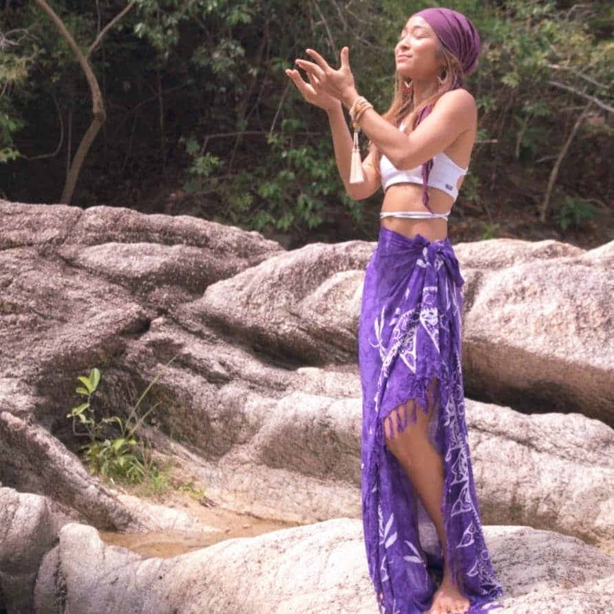 Woman standing on rocks in nature with her hands out to nature in reverence wearing a head scarf, white top and purple sarong as a skirt. 