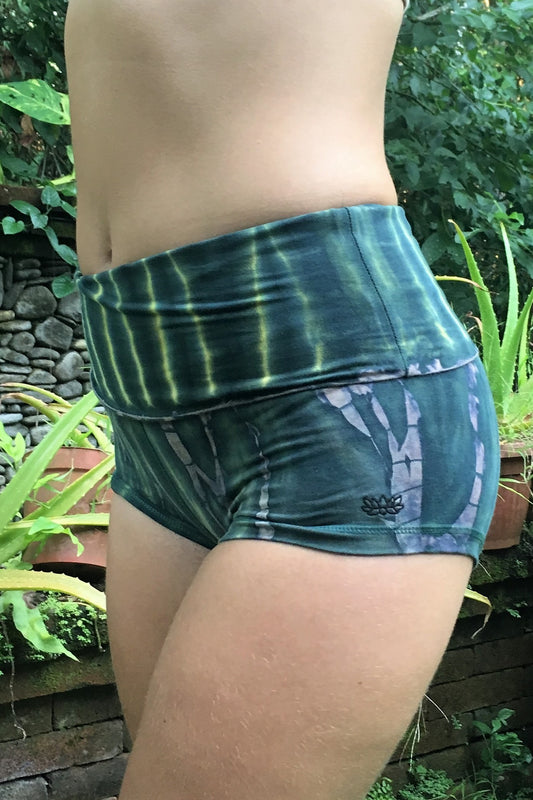 Side view of Cotton RainForest yoga shorts tie dyed in green hues with beige highlights and small lotus flower embroidery on side.